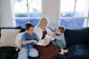 A woman sits on a dark sofa holding a newborn baby while two young boys sit beside her near bright windows.