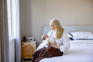 A blonde mother sits on a bed cradling her newborn baby, wrapped in a brown blanket, in a serene bedroom.