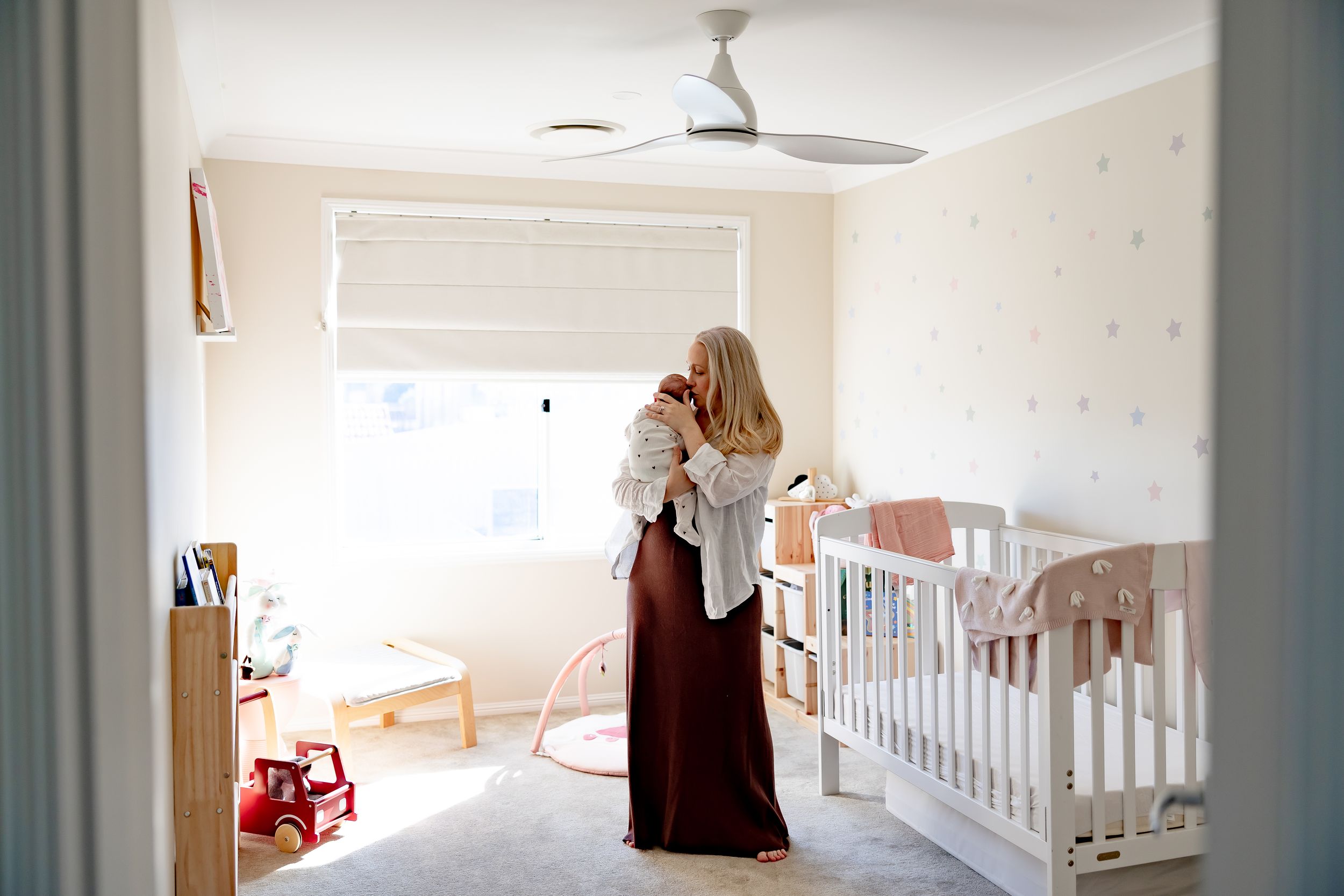Mother holding newborn baby in a softly decorated nursery room with white crib and pastel star wall decals.