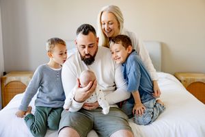 Happy family of five with newborn baby sitting together on white bed smiling.