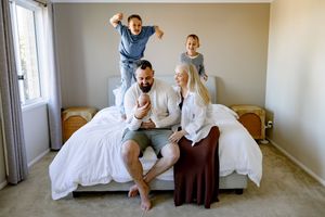Happy family with two kids jumping on bed, parents holding newborn baby in bright bedroom.