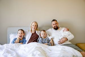 Happy family of five with two boys and a newborn baby posing together on a white bed at home.