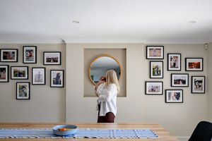 Woman arranging family photo gallery wall with framed pictures around round mirror at home.