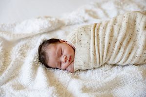 Peaceful newborn baby swaddled in a cream speckled blanket, sleeping on a soft white fluffy surface.
