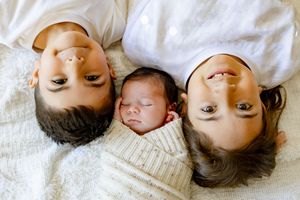 Two parents and a sleeping newborn baby lying together on a white bed, captured in a tender family portrait.