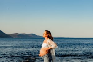 Silhouette of pregnant woman against ocean sunset with mountains in distance.