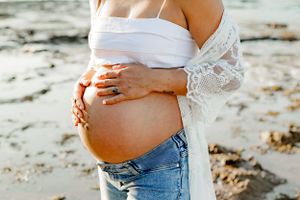 Side view of pregnant belly in jeans and white lace wrap on sandy beach at sunset.