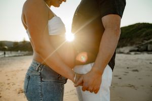Intimate sunset maternity photo with couple holding hands on beach with sun flare.