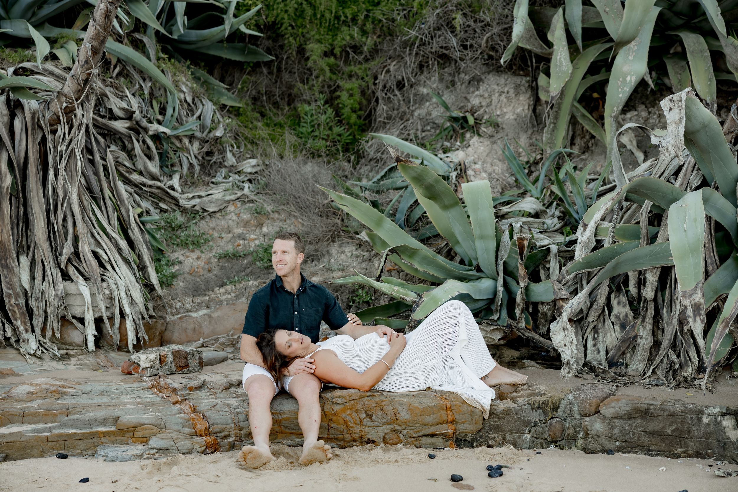 A couple rests on rocky terrain surrounded by large coastal plants.