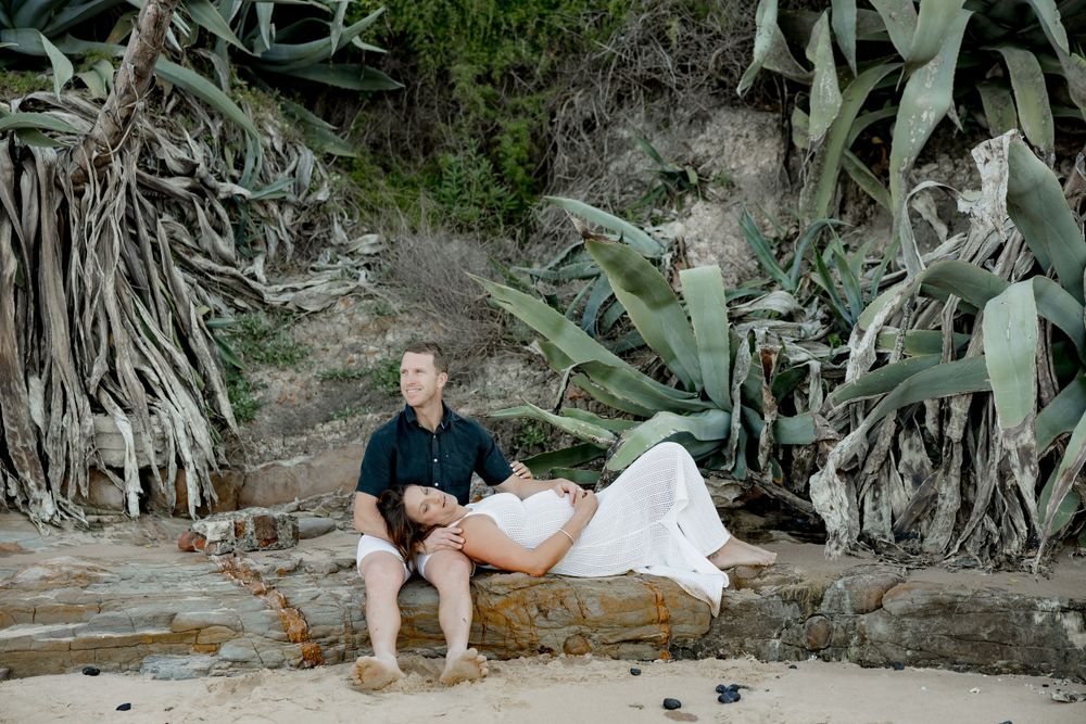 A couple rests on rocky terrain surrounded by large coastal plants.