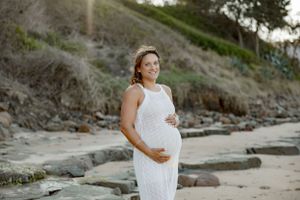 A maternity photoshoot on a rocky beach featuring a white lace dress against a coastal backdrop at sunset.
