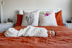 A baby wrapped in white swaddle blanket lies on a rust-colored bed with decorative pillows.