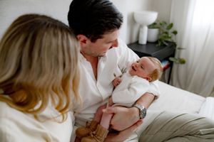 A newborn baby wrapped in white is cradled in a cozy bedroom setting with soft natural lighting.