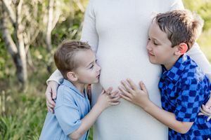Two young boys lean against their pregnant mothers belly outdoors.