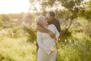 Couple embracing outdoors at golden hour, pregnant woman in white dress with partner kissing her head in lush green field.