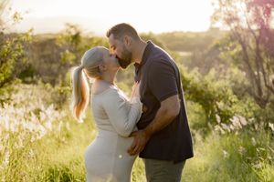 Couple sharing a romantic kiss outdoors in a sunlit meadow, woman in white dress with blonde ponytail, golden hour.