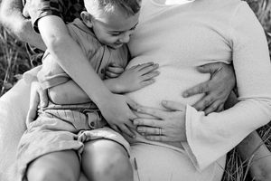 Black and white maternity photo of pregnant woman with child kissing baby bump outdoors.