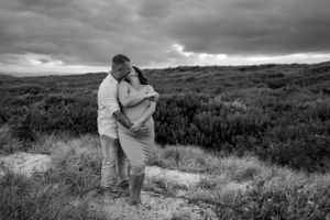 Black and white photo of couple embracing on hillside with dramatic cloudy sky.