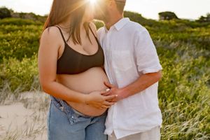 Intimate pregnancy photoshoot at sunset on a beach with couple embracing among dunes and grass.