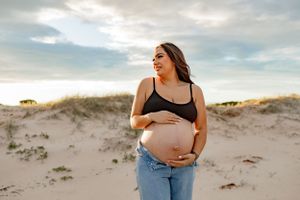 Expecting mother in casual attire poses on sandy dunes during golden hour photoshoot.