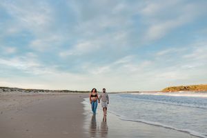 Couple walking together along ocean shoreline during sunset beach maternity session.