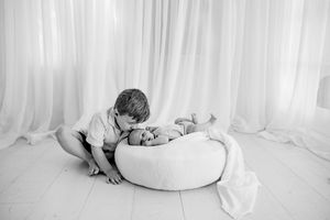 Black and white photo of a young boy leaning over a round white bean bag to gently kiss a sleeping newborn baby.