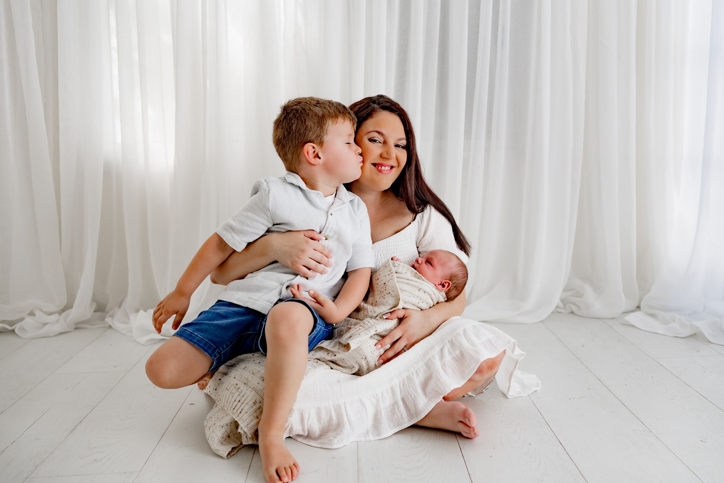 A mother sits on white floor holding newborn while young son kisses her cheek in bright studio setting.