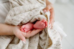 Tiny newborn baby feet cradled in mother's hands, wrapped in a soft cream knit blanket.