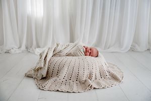 Newborn baby sleeping peacefully wrapped in a cream crochet blanket on a white studio floor.