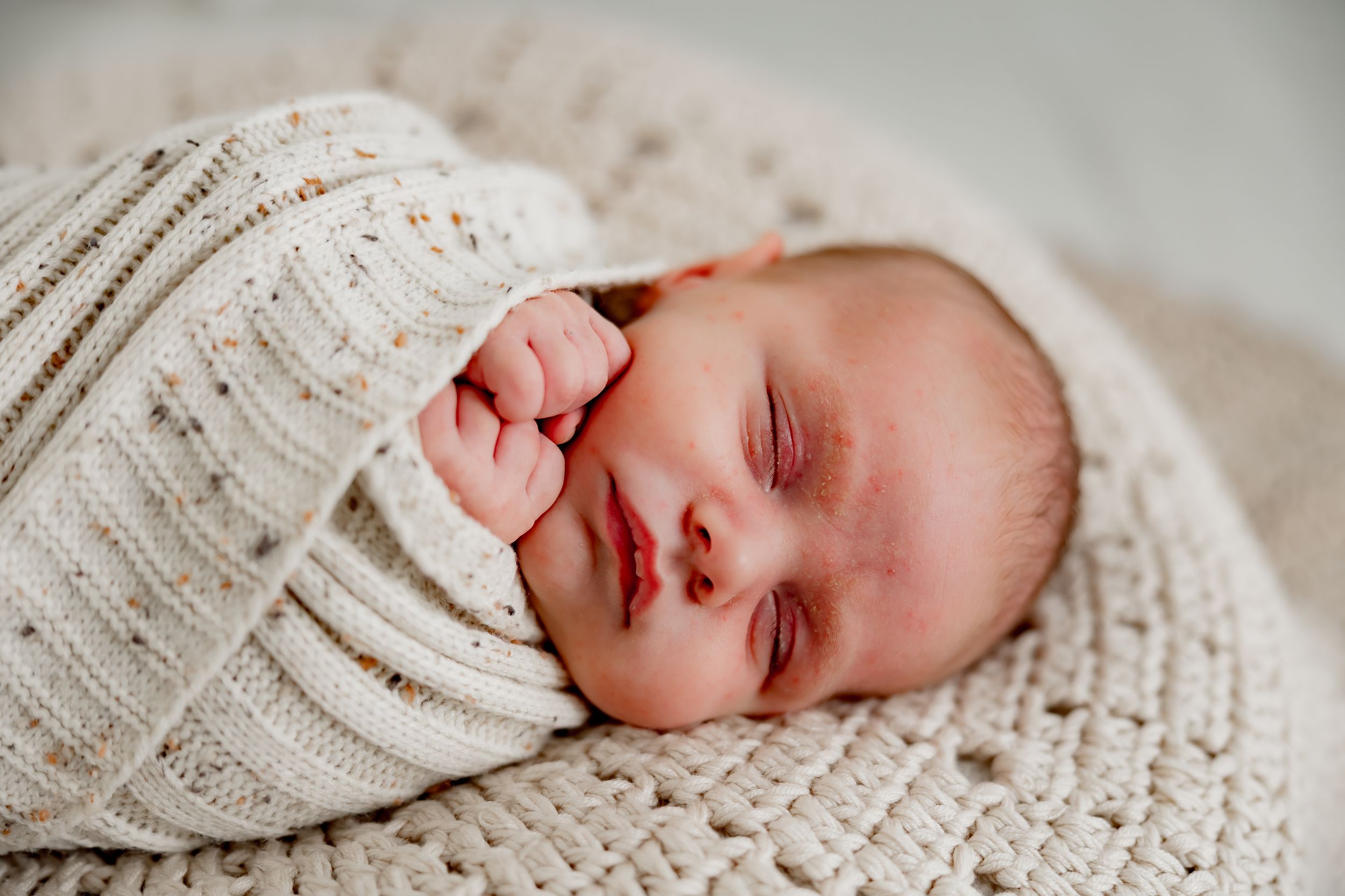 Peaceful newborn baby swaddled in a cream knit blanket, sleeping with tiny fist tucked under cheek.