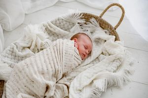 Newborn baby sleeping peacefully swaddled in white knit blanket inside a woven Moses basket.
