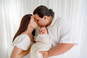 New parents kissing over their swaddled newborn baby, dressed in white against a soft white curtain backdrop.