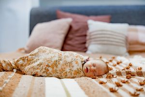A baby wrapped in a floral swaddle blanket rests on a striped bed with brown and rust colored throw pillows.