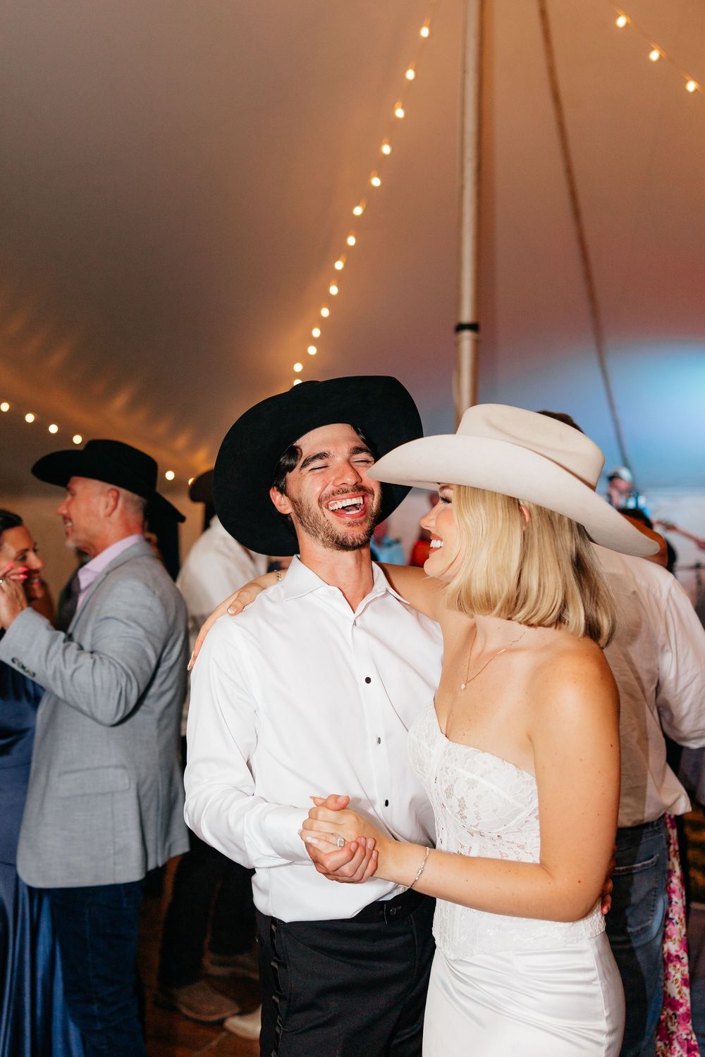 Couples dancing and embracing under twinkling lights at a wedding reception in a tent venue.
