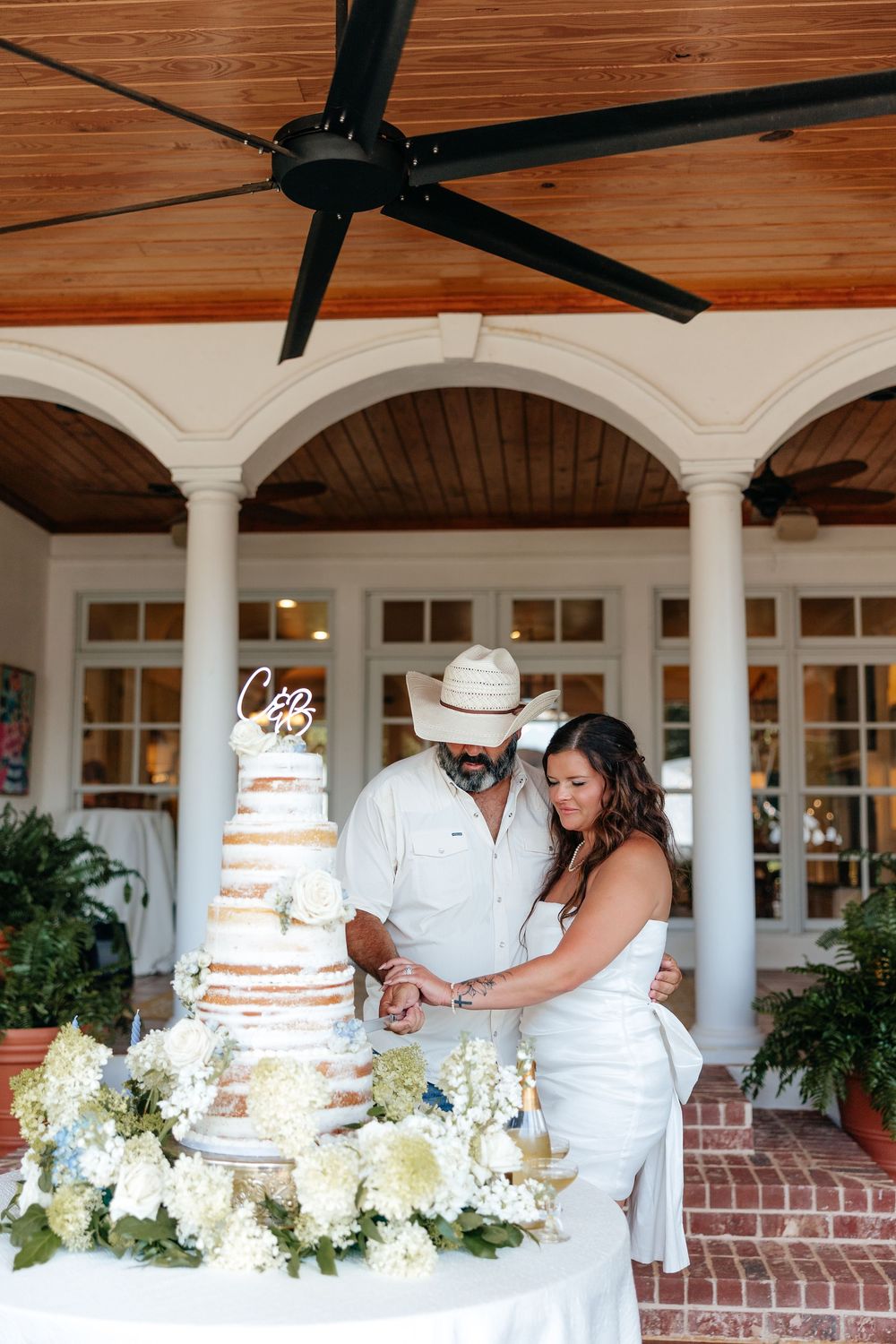 Couple cutting a tiered naked wedding cake adorned with white florals outdoors under a covered patio with ceiling fan.