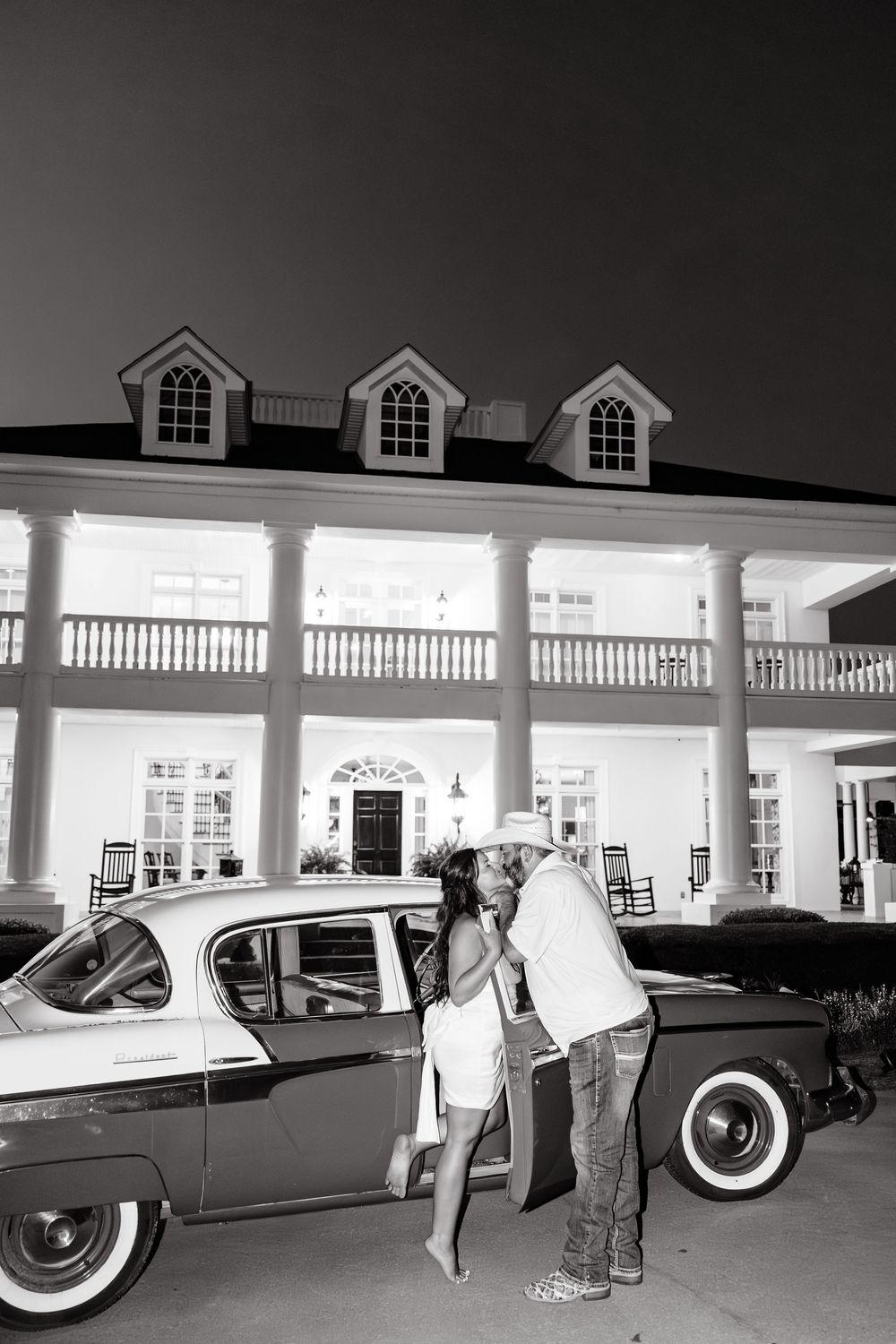 Couple kissing beside a vintage classic car in front of an illuminated colonial mansion at night, black and white photo.