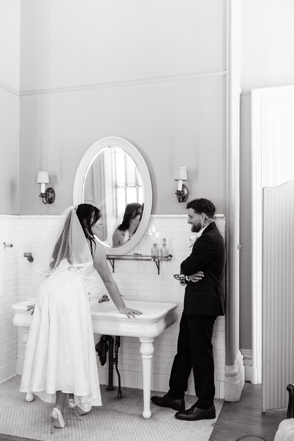 A bride in a white dress and groom in a suit share a candid moment in an elegant vintage bathroom with pedestal sink.