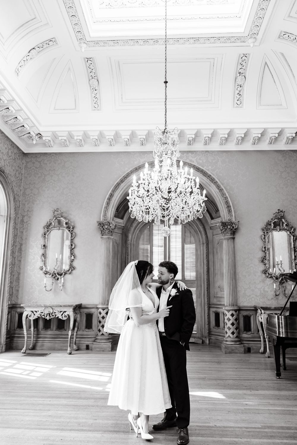 A couple shares an intimate moment in an elegant ballroom with ornate moldings and a crystal chandelier.