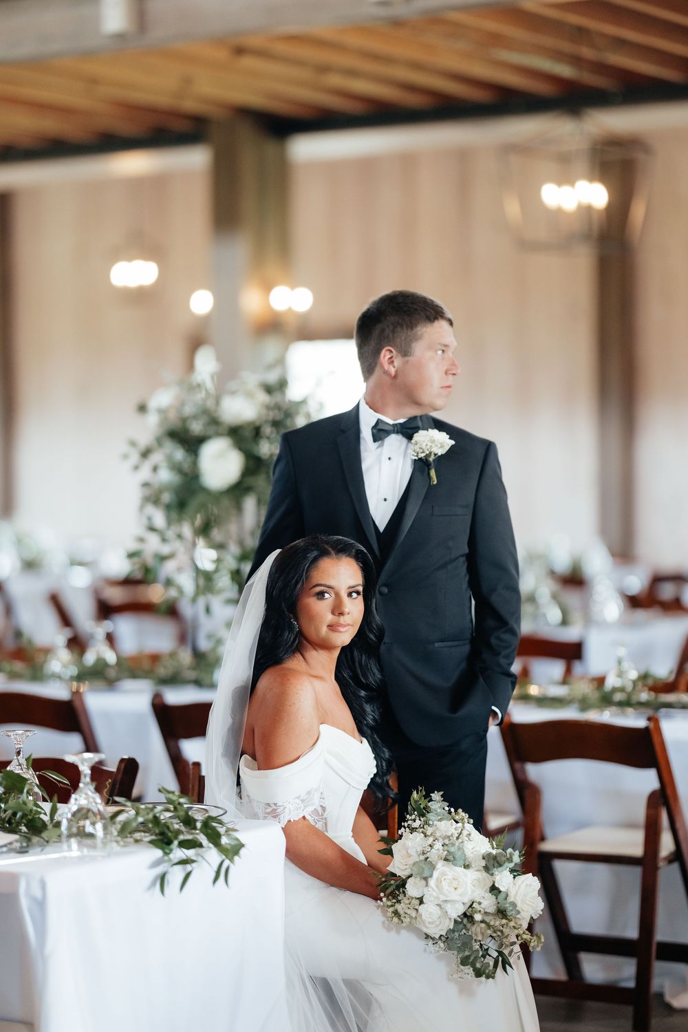 Elegant wedding couple poses in reception venue with white floral decorations and wooden chairs.
