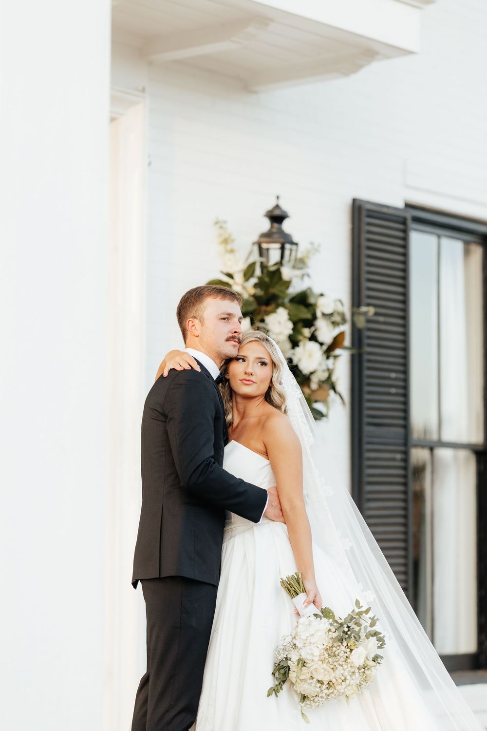 Bride in strapless white ballgown holding bouquet embraces groom in black tuxedo outside elegant white building.