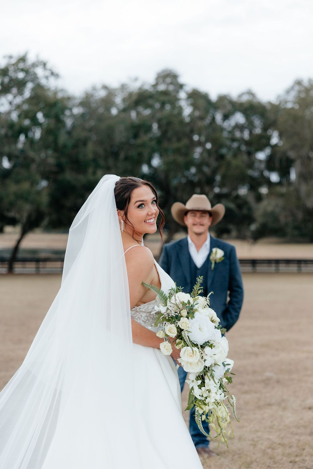 Bride in white gown and veil holds bouquet, smiling, with groom in navy suit and cowboy hat in background.