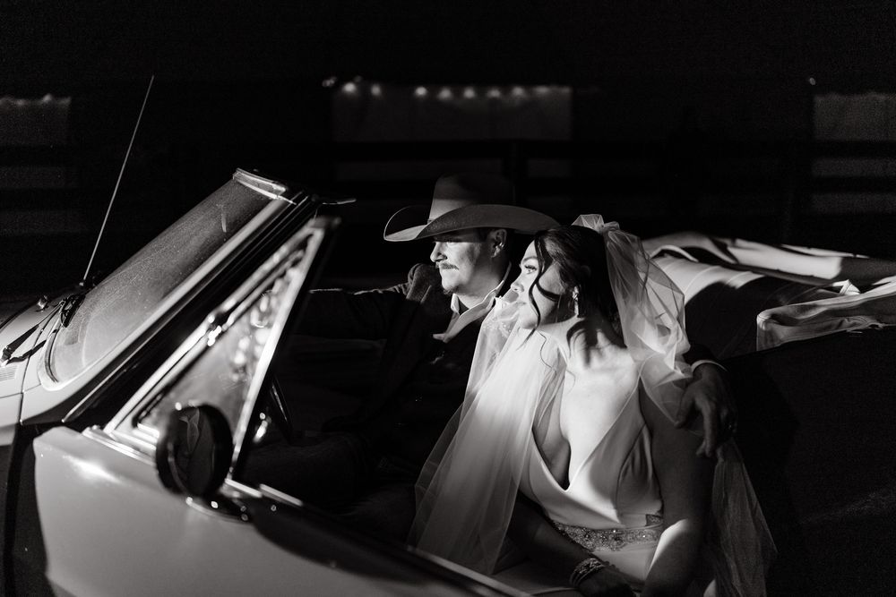Black and white photo of a bride and groom in a convertible car, groom wearing cowboy hat, bride in veil.