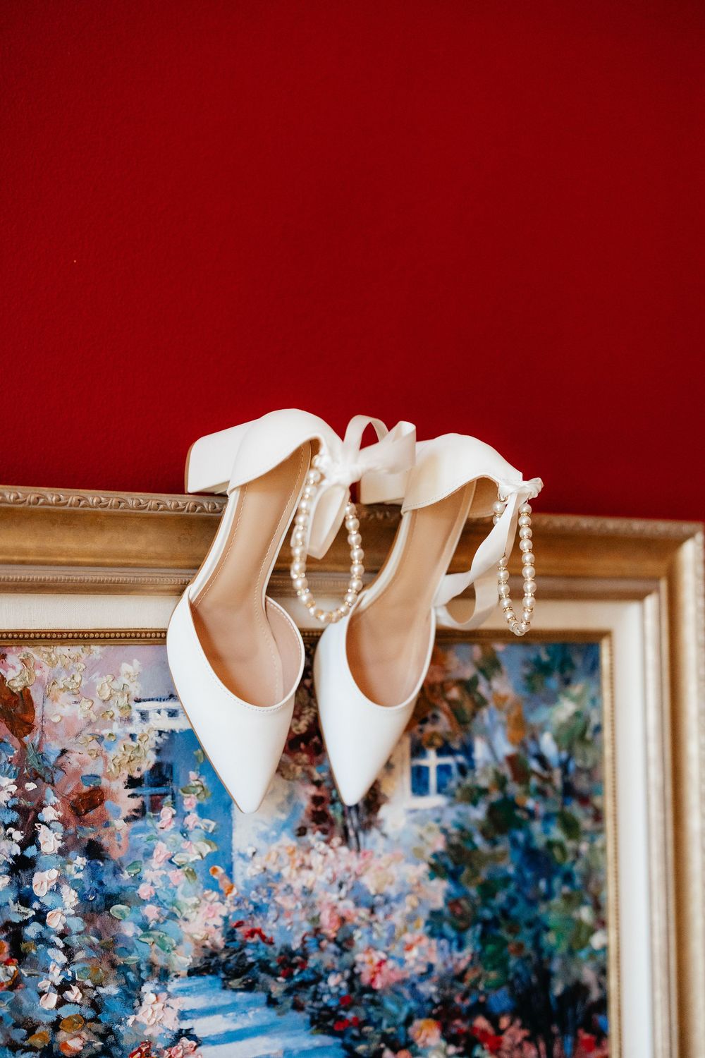 White bridal heels with pearl ankle straps and satin ribbons hanging on a gold picture frame against a red wall.