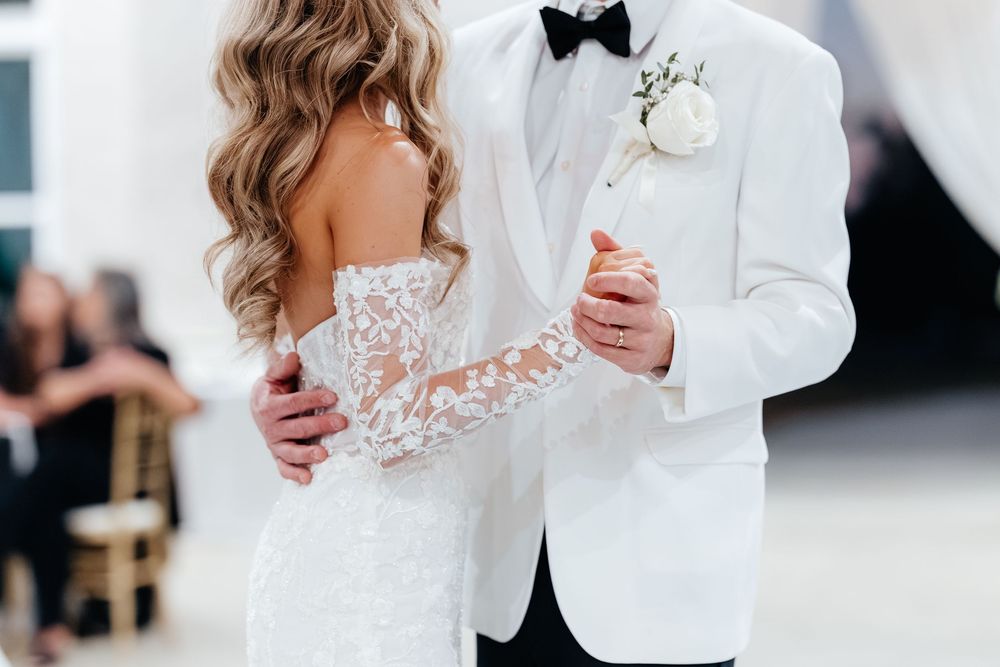 Bride and groom share first dance, bride in lace gown, groom in white tuxedo with boutonniere.
