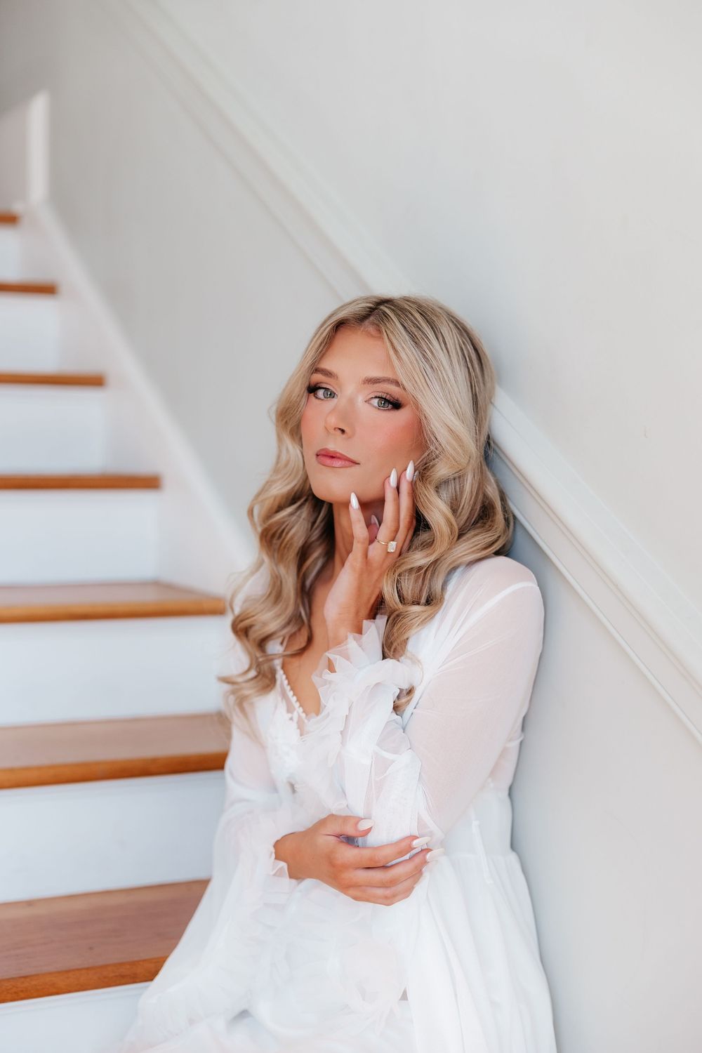 Blonde bride in white lace robe sitting on staircase steps, posing gracefully with hand near face.