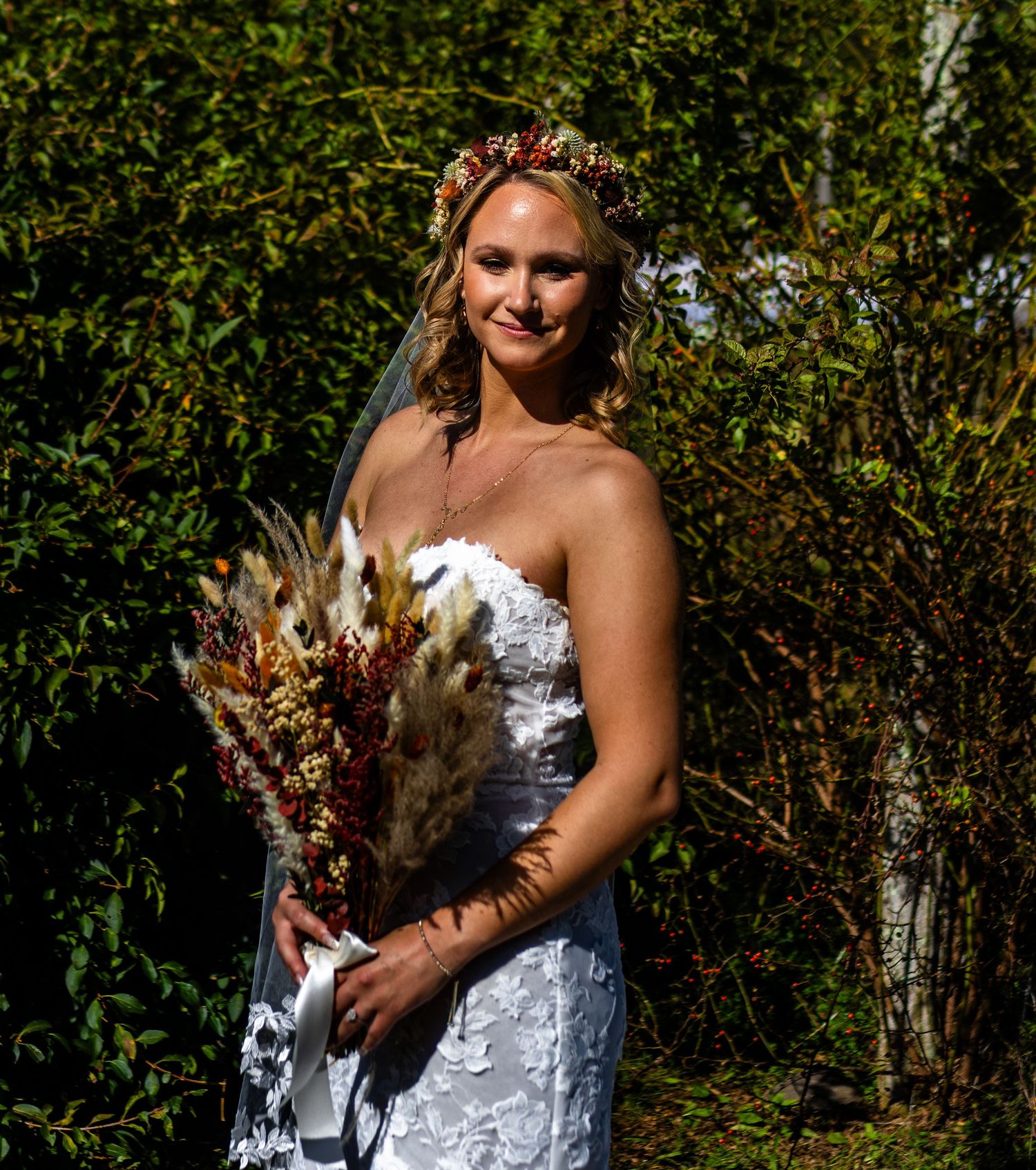 A bride in a white lace wedding dress holds a rustic autumn-themed bouquet with dried flowers and grasses.