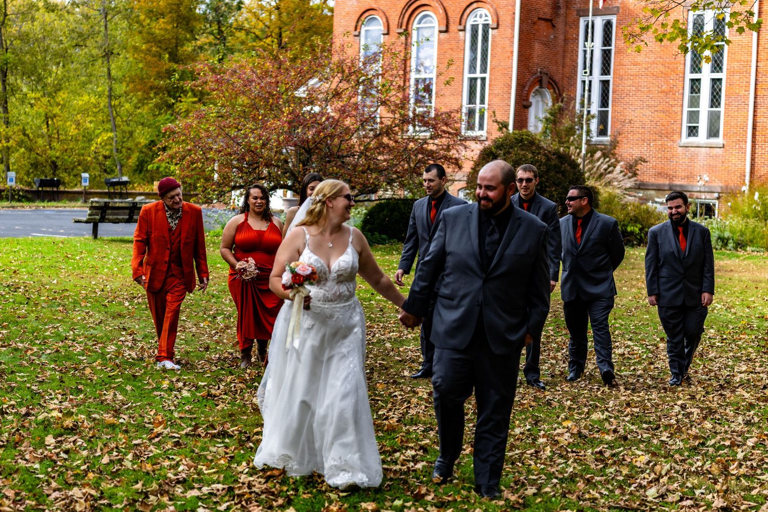 Wedding party poses in front of historic red brick building with autumn foliage in the background.