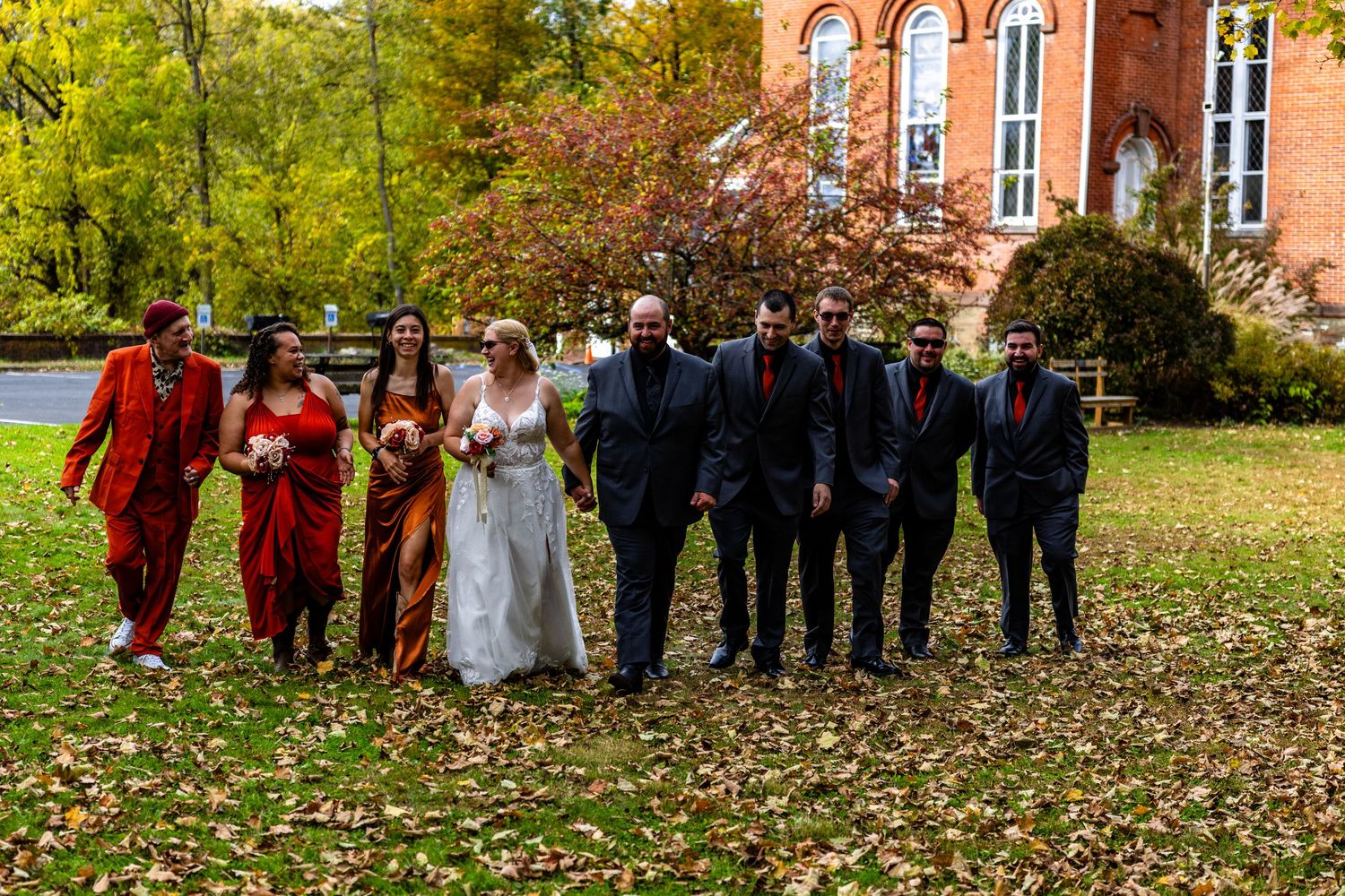 A wedding party stands in line outdoors during autumn with colorful fall foliage and a brick building in the background.