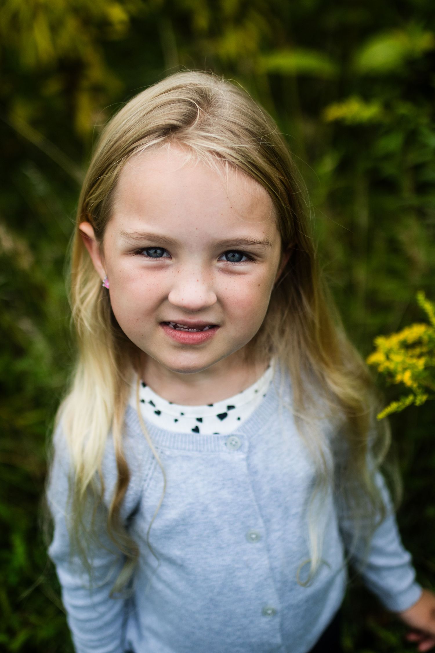 Young child in a light gray cardigan standing against a green garden background.