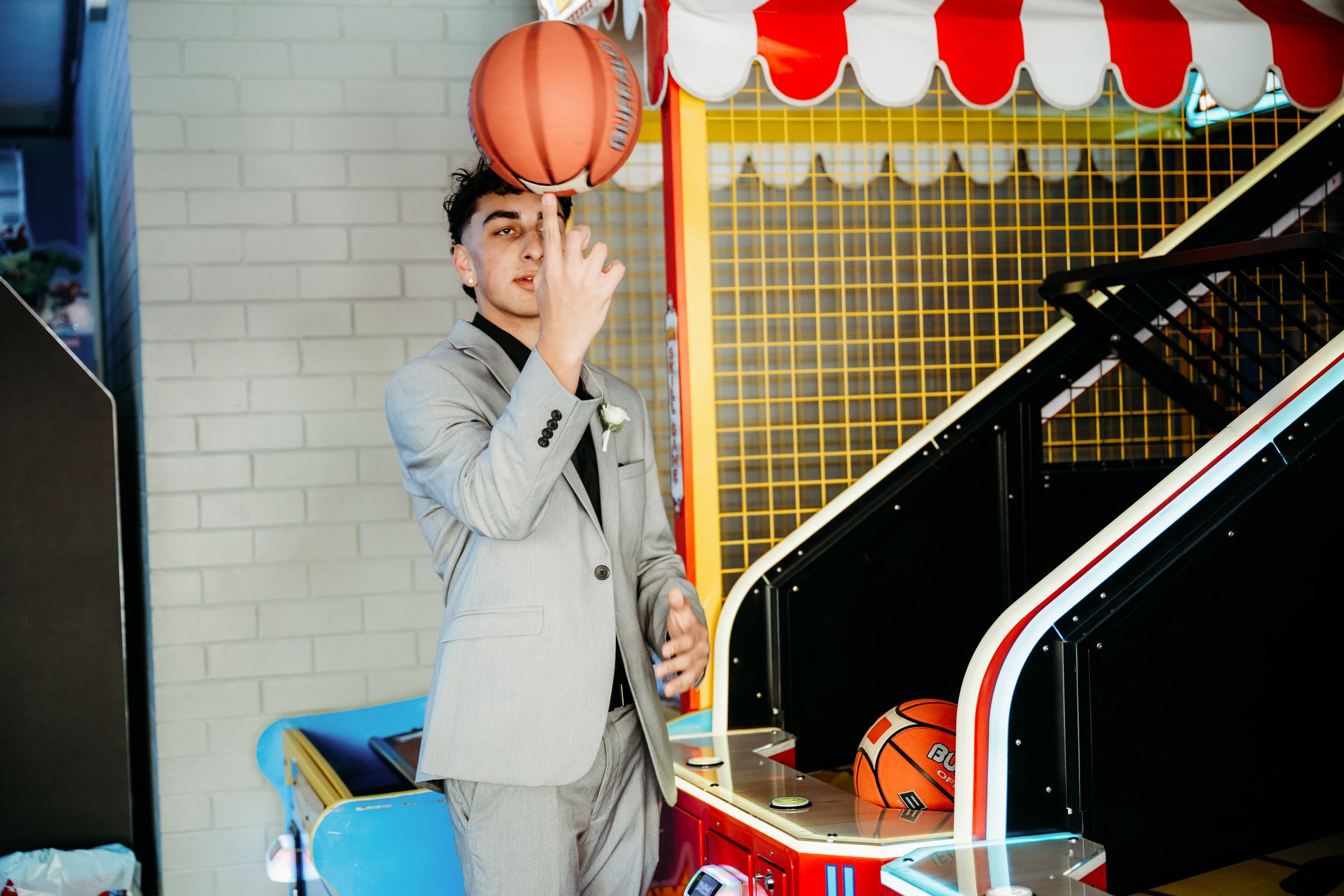 A person in a gray suit poses playfully with a basketball near an arcade or game area with red and white decor.